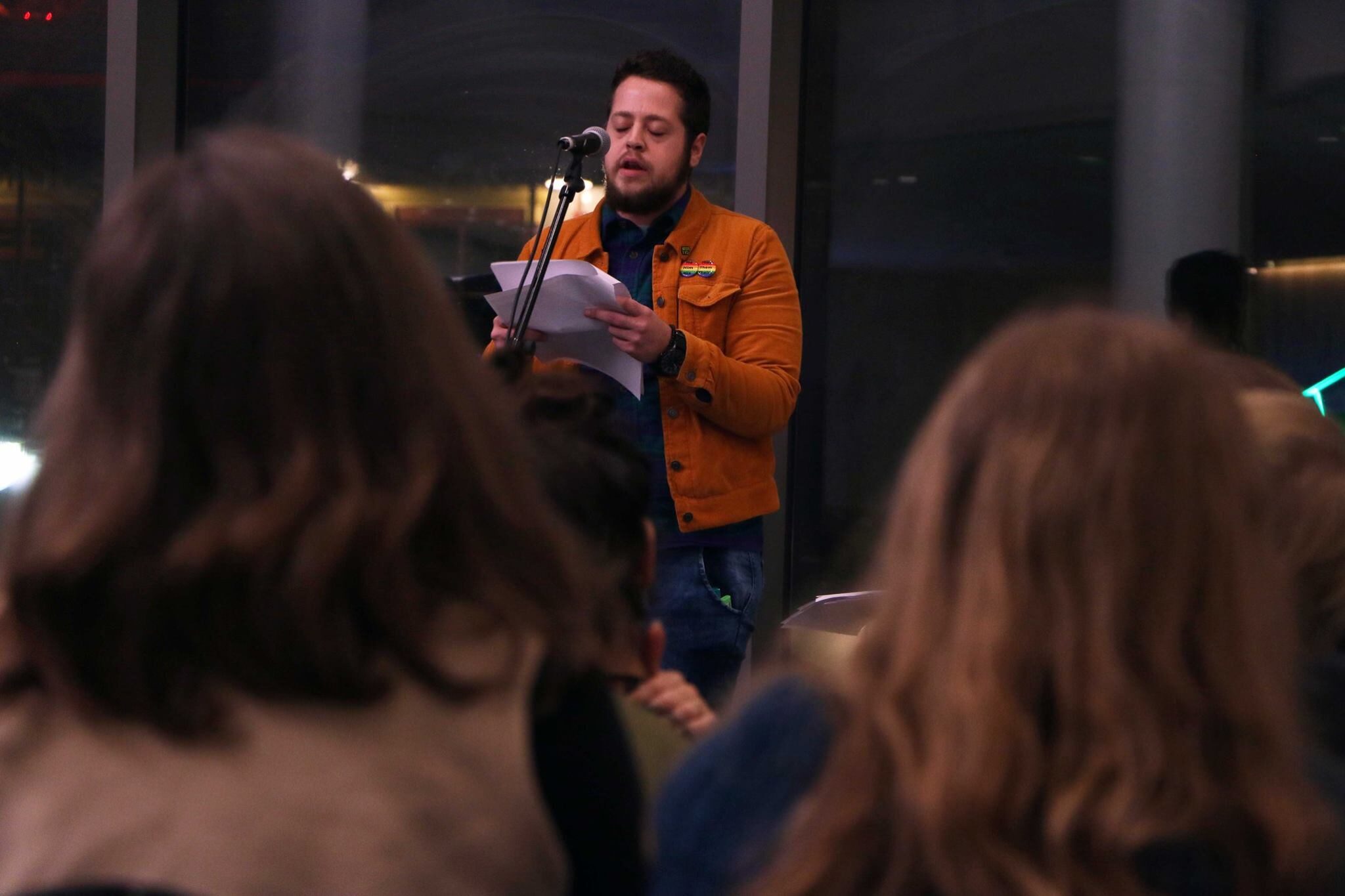 The poet Brody Parrish Craig performs at an in-person event. They are on a stage with a microphone, and they hold pieces of paper in their hands. They have short brown hair and are wearing an orange jacket.